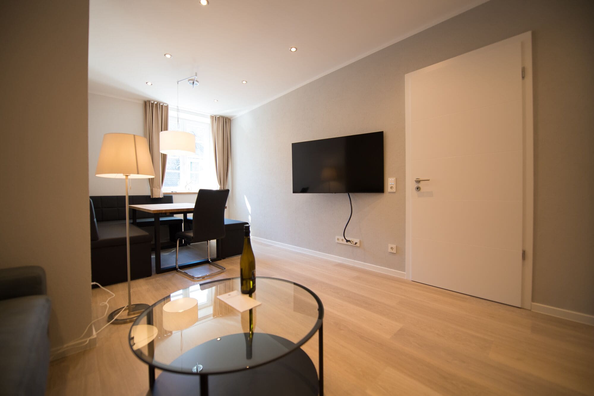 A modern living room with a flat-screen TV mounted on the wall, a glass coffee table, a sofa, a dining table with chairs, and a lamp.