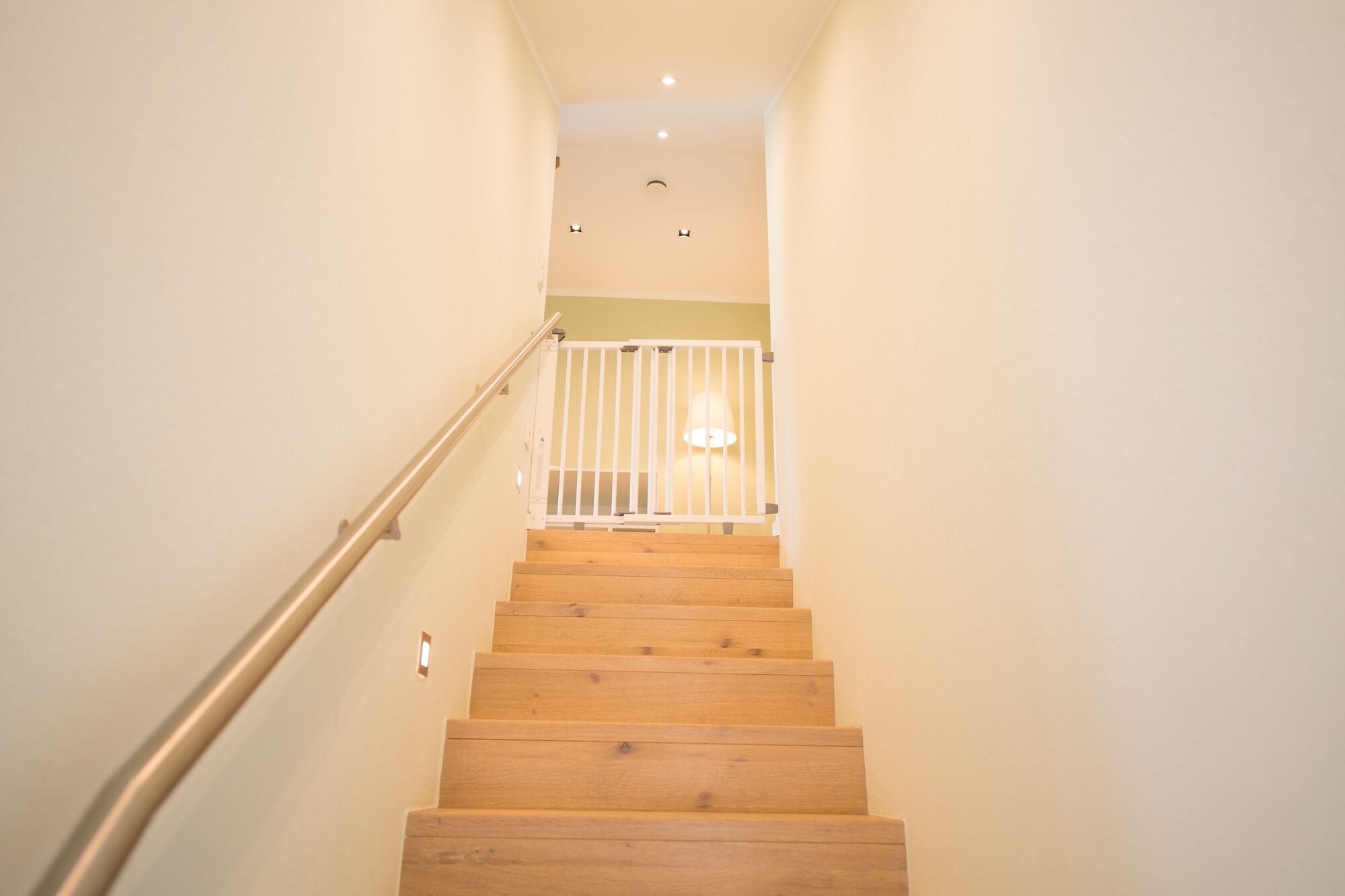 A wooden staircase with a metal handrail leading to a landing with a white railing and a lamp.