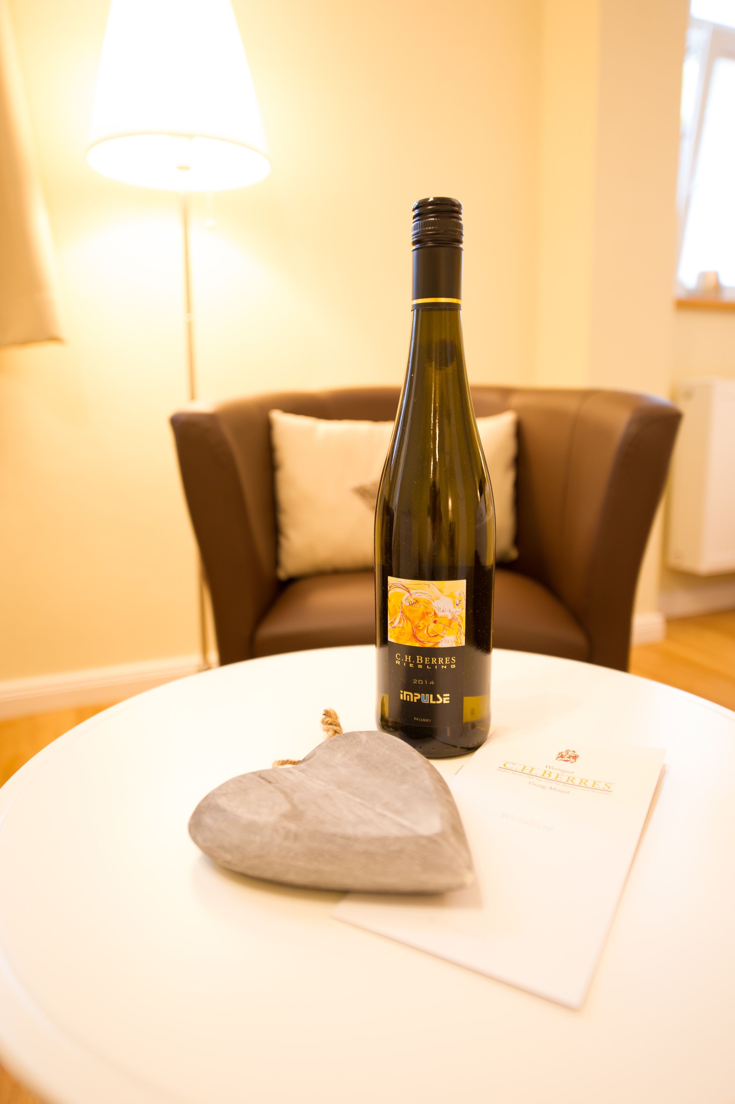 Uma garrafa de vinho sobre uma mesa branca, com um almofado e um menu em um quarto com um sofá castanho e uma luminária ao fundo.