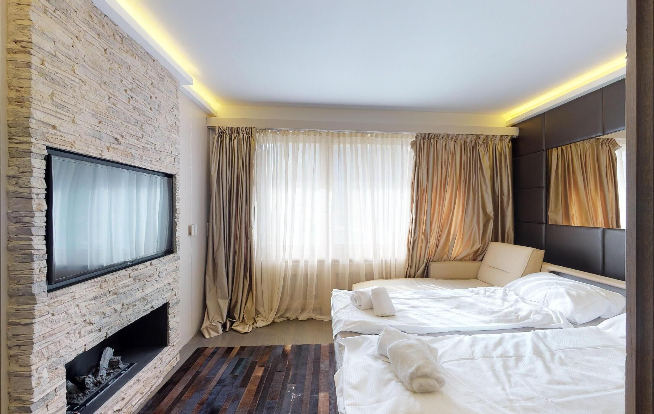Une chambre d’hôtel avec une cheminée en pierre, un lit avec du linge de maison blanc, un canapé et un téléviseur fixé au mur.