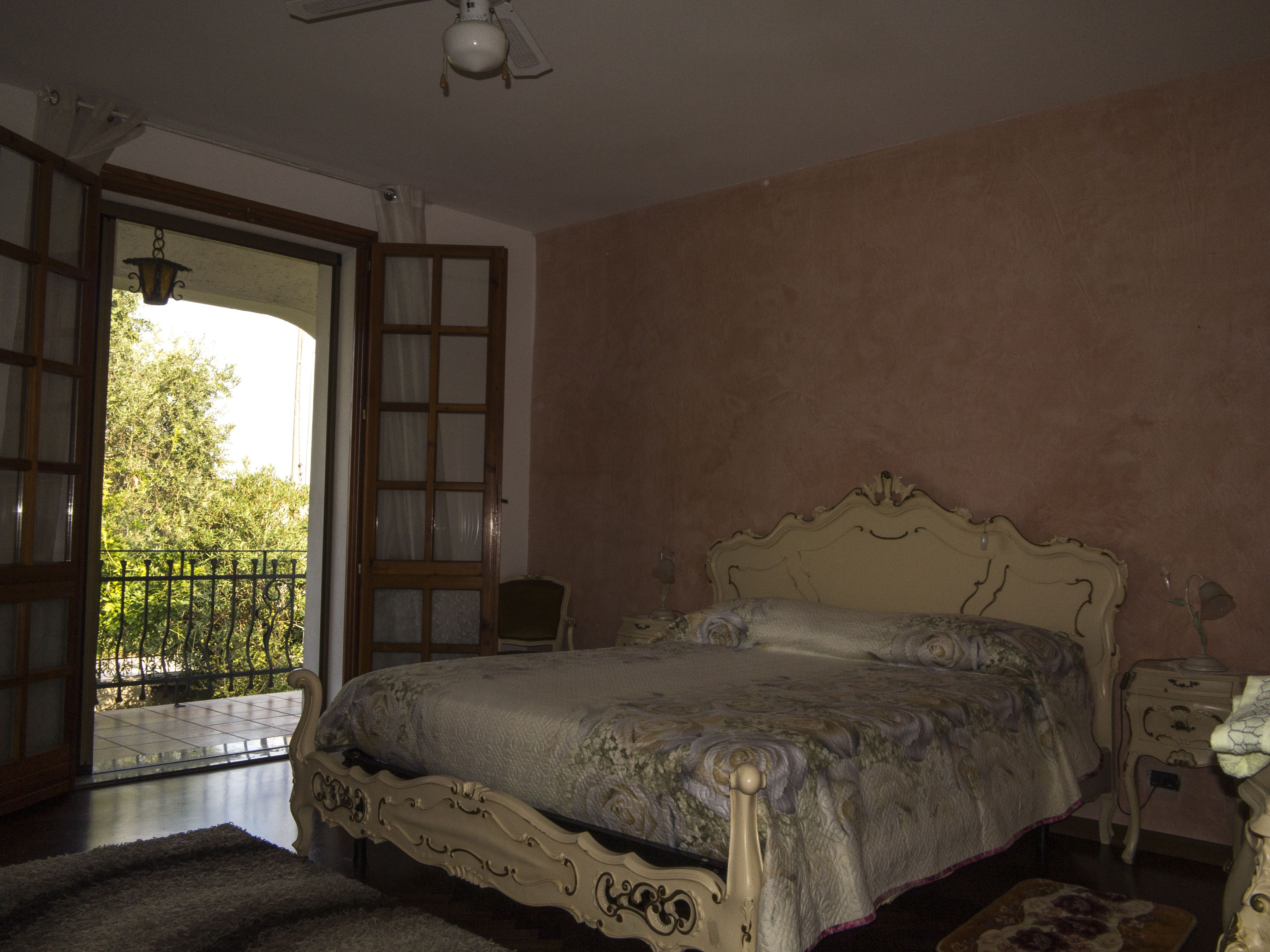 A bedroom with a large bed, a nightstand, and a chair. There is a balcony with a railing and a view of trees.