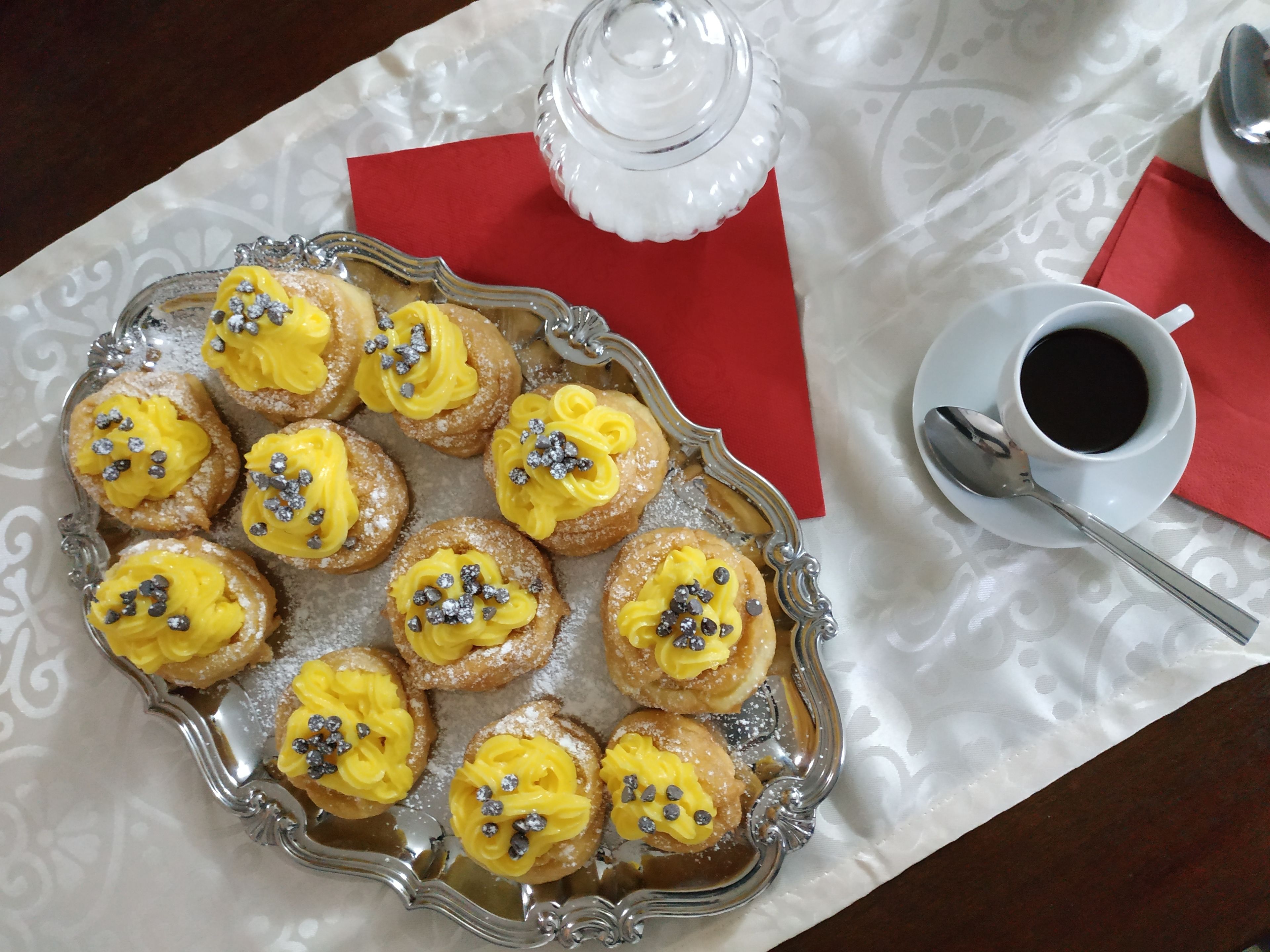 Une assiette de pâtisseries à la crème jaune et aux décorations argentées, un récipient en verre, une tasse de café et une cuillère, le tout posé sur une table recouverte d’une nappe blanche et agrémentée de serviettes rouges.