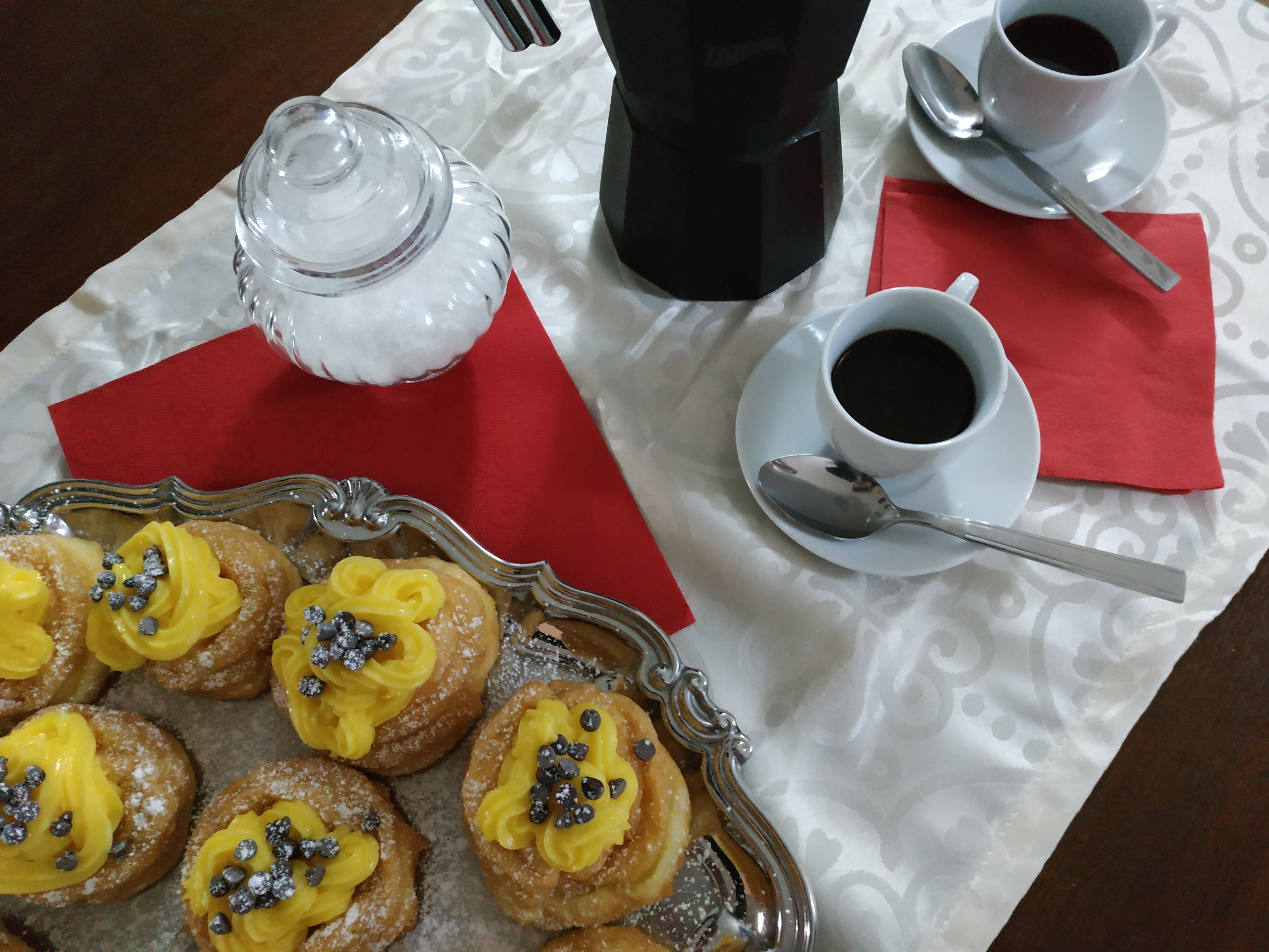 En bakke med wienerbrød, gul creme og chokoladestykker, en kaffemaskine, to kopper kaffe og en sukkerdåse på et bord.