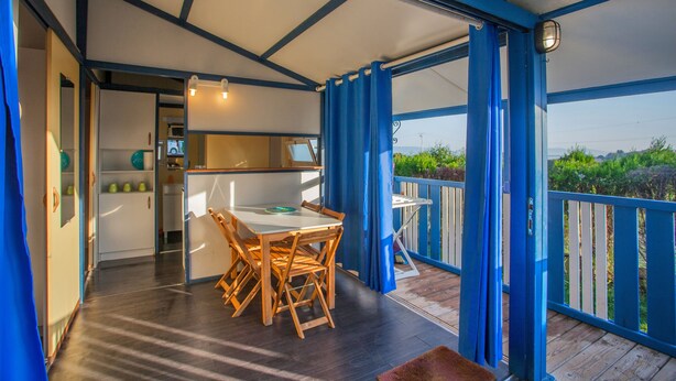 A covered outdoor dining area with a table and chairs, a view of the outdoors, and a kitchen area in the background. - Camping Os Fieitás (O Grove)