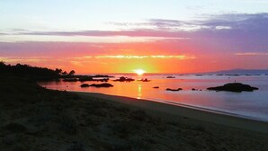 A beach at sunset with a clear sky, calm water, and scattered rocks. - Camping Os Fieitás (O Grove)