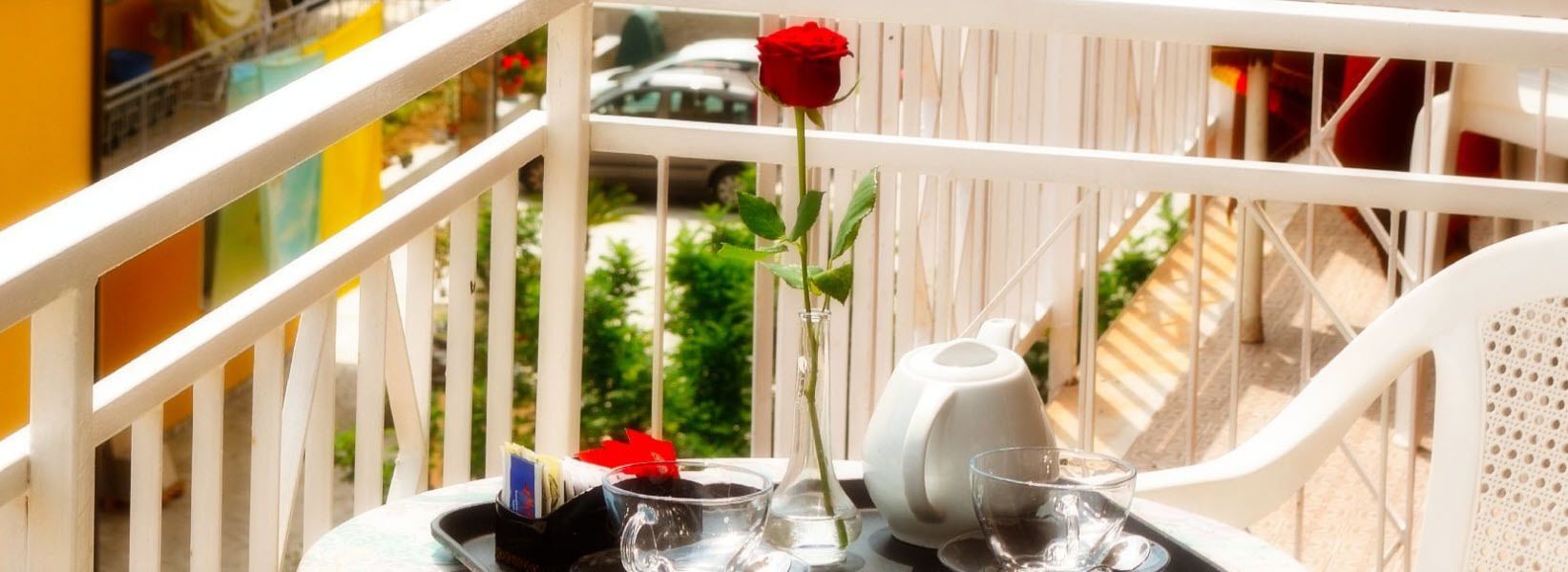 A balcony with a white railing, a table with a teapot, a glass, and a red rose.