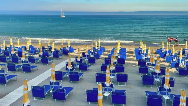 A beach with rows of blue chairs and yellow umbrellas, a sailboat in the distance, and a clear sky. - Hotel Jole (Andora)