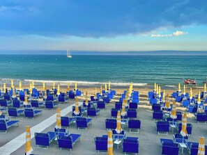 Une plage bordée de rangées de chaises bleues et de parasols jaunes, un voilier à l’horizon et un ciel dégagé.