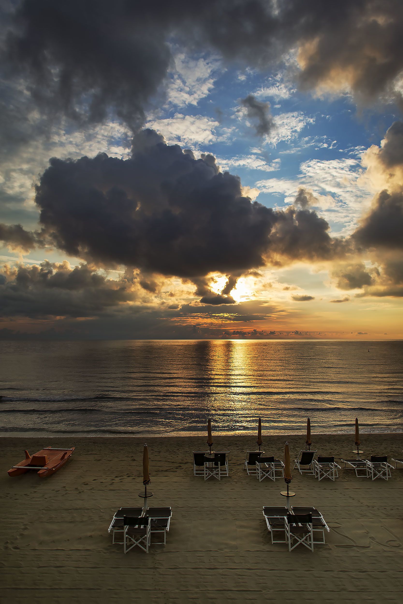 A beach with rows of sun loungers and umbrellas, a boat, and a sunset over the ocean.
