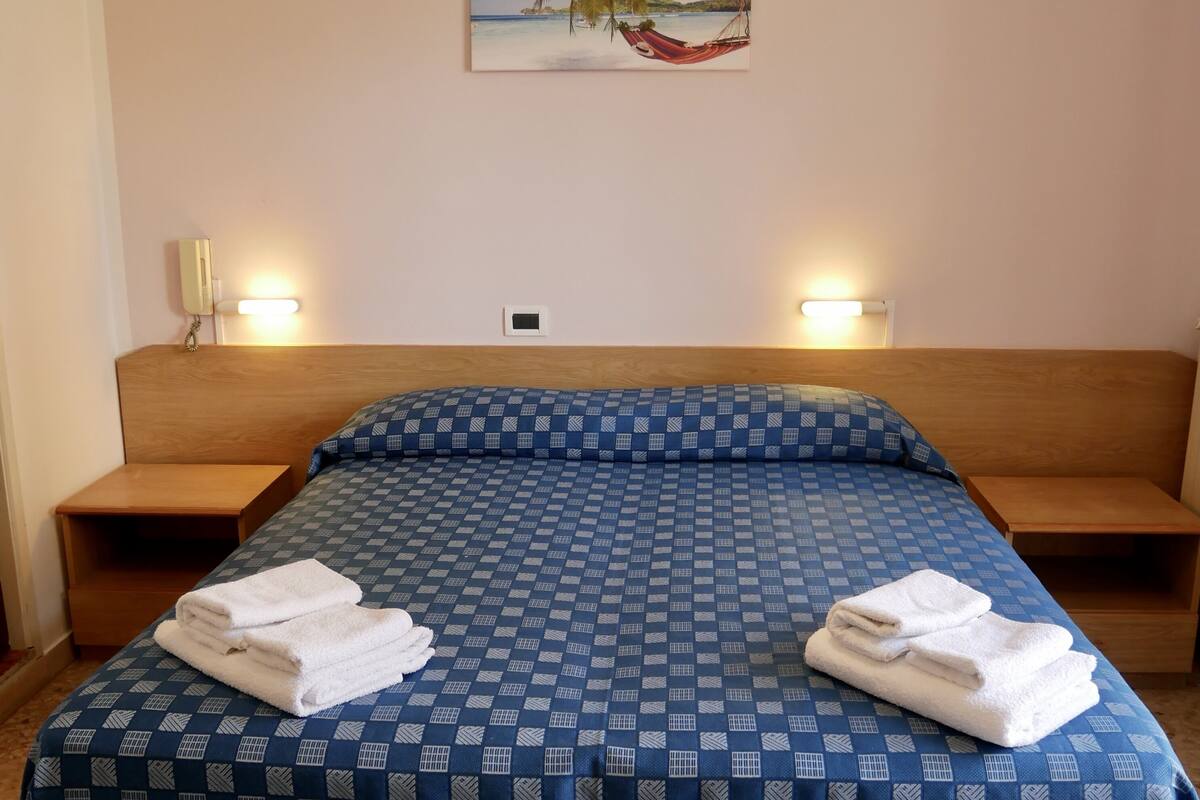 Una cama bien hecha con una colcha azul a cuadros, dos mesitas de noche con lámparas, y un cuadro de una playa en la pared.