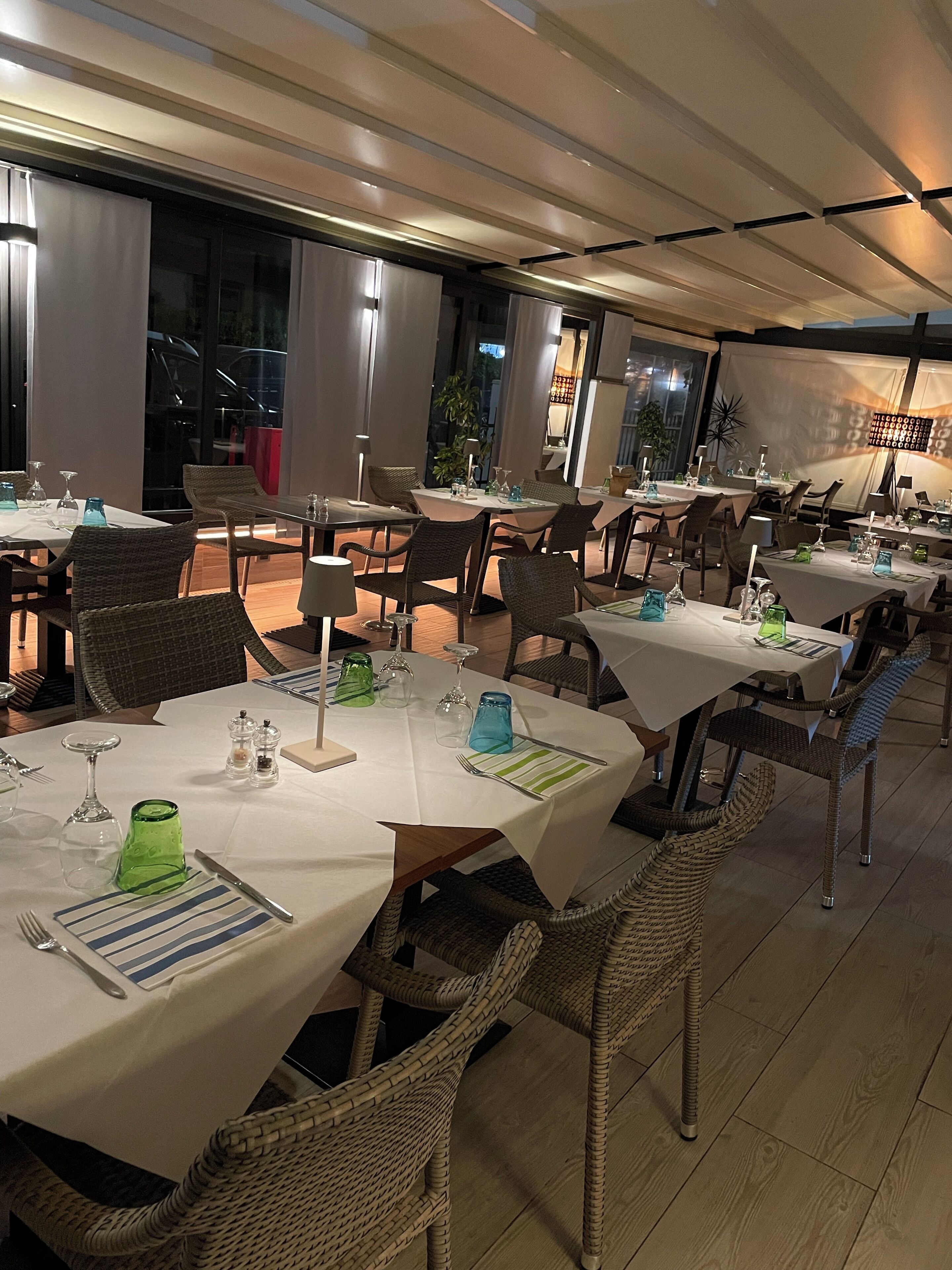 Restaurante com várias mesas montadas para refeições, com cadeiras de vime e toalhas de mesa brancas.