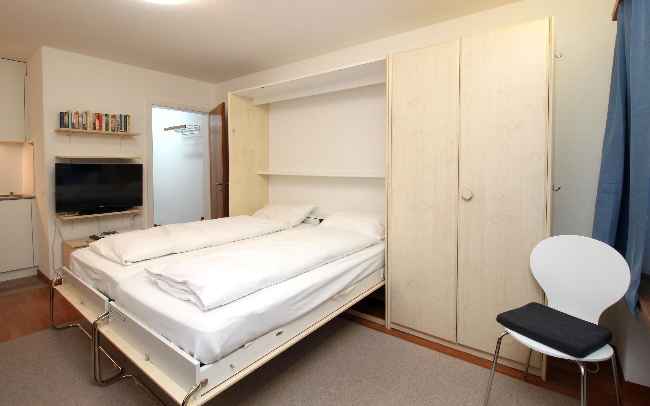 Ein gemütliches Schlafzimmer mit Bett, Fernseher, Bücherregal und Kleiderschrank.
