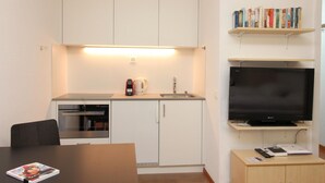 A compact kitchenette with a built-in oven, a coffee maker, and a sink. There is a flat-screen TV mounted on the wall above a wooden shelf with books. A dining table with a magazine is in the foreground. - Popular and centrally located residential building in St. (St. Moritz)