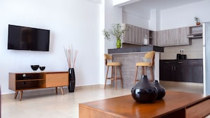 A modern living room with a flat-screen TV, wooden furniture, and a kitchen area in the background. - L'Aziz Suites (Nairobi)