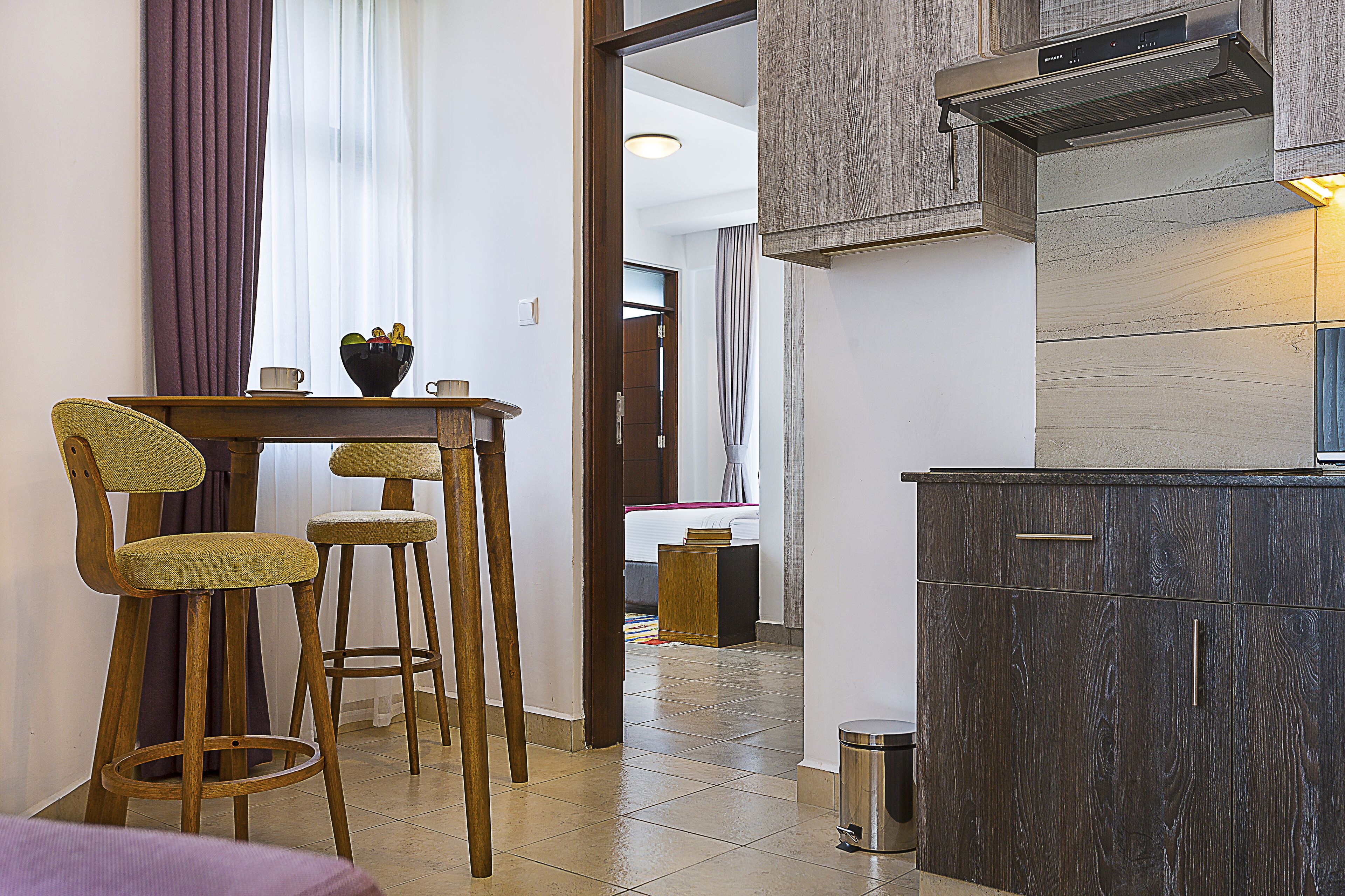 A compact kitchen with dark wood cabinets, a stainless steel range hood, and a small dining area with two stools and a table.