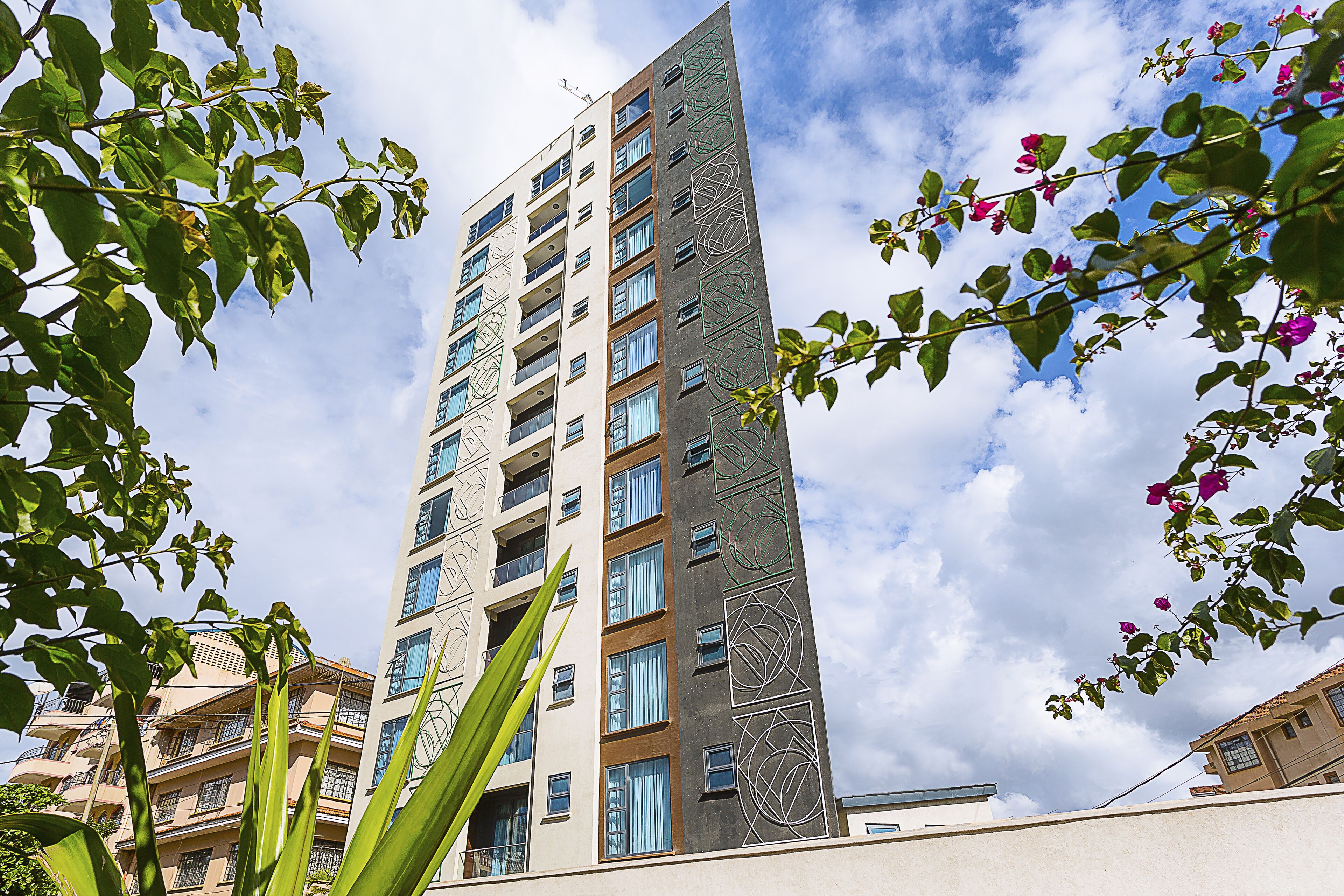 A tall, modern building with multiple balconies and windows, surrounded by greenery and flowers.