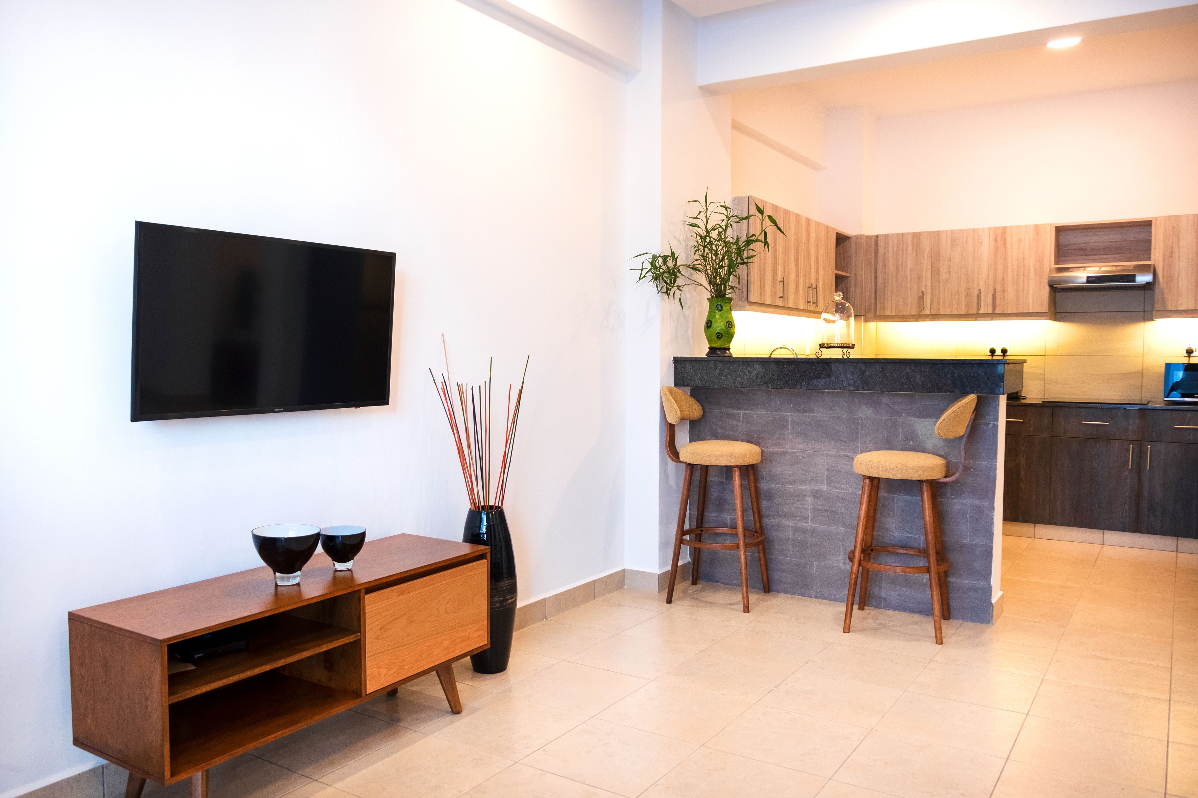 Una cocina moderna con armarios de madera, encimera negra y estufa integrada. Hay un televisor montado en la pared, una mesa consola de madera y dos taburetes de bar.