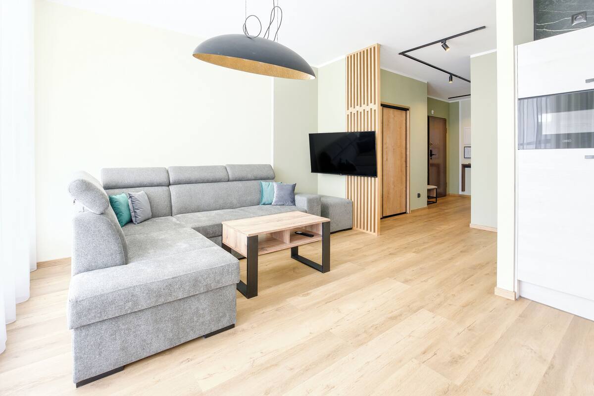 A modern living room with a grey sectional sofa, a wooden coffee table, a TV mounted on a wooden panel, and a kitchen area with white cabinets.