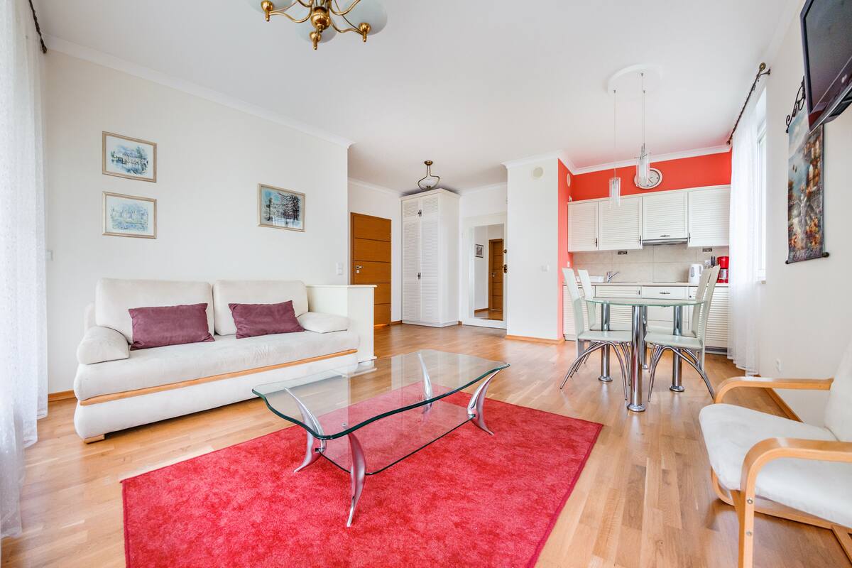 A modern living room with a glass coffee table, a red rug, a white sofa, and a dining area with a glass table and chairs.