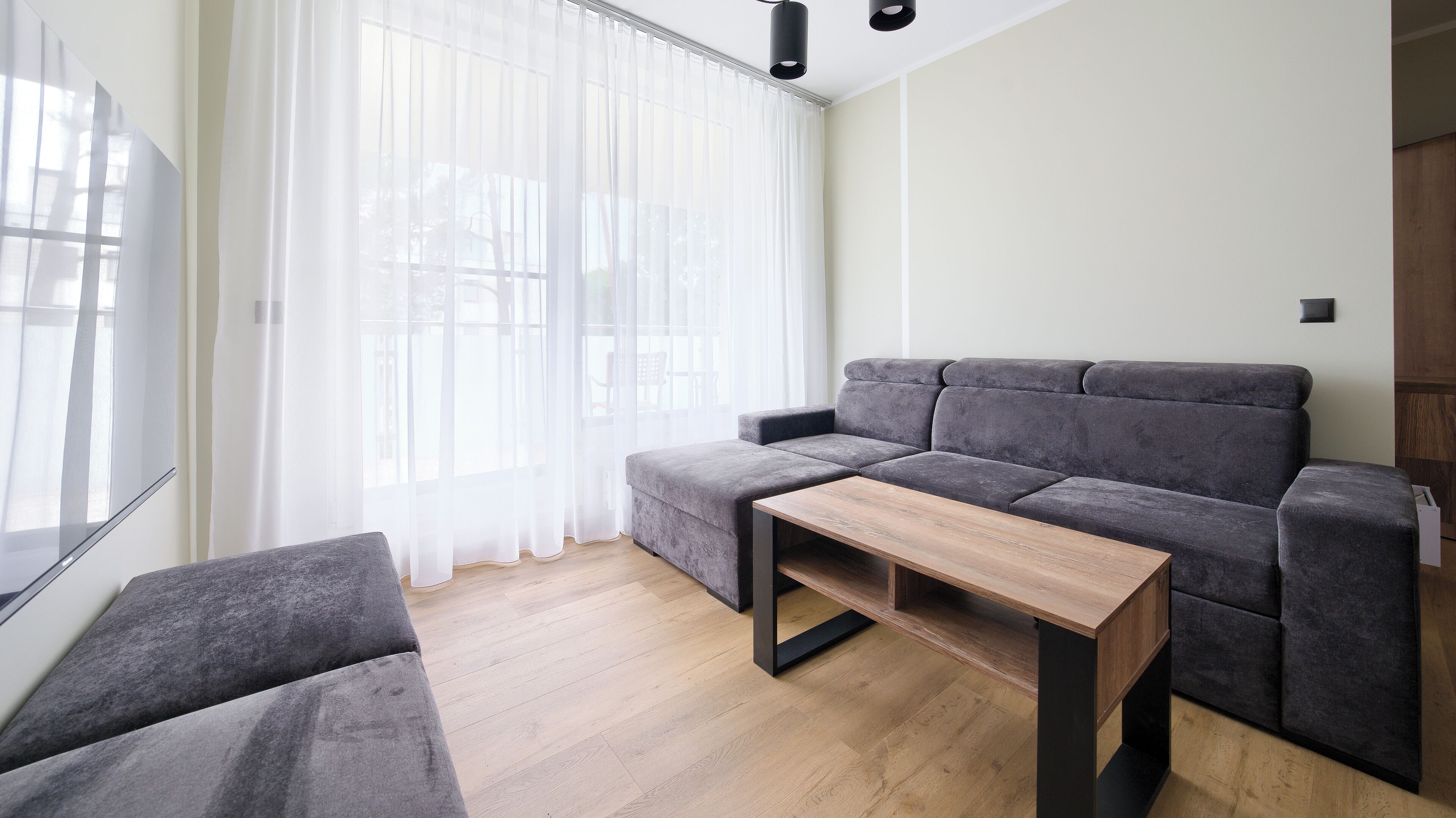 A modern living room with a grey sofa, a wooden coffee table, and sheer white curtains.