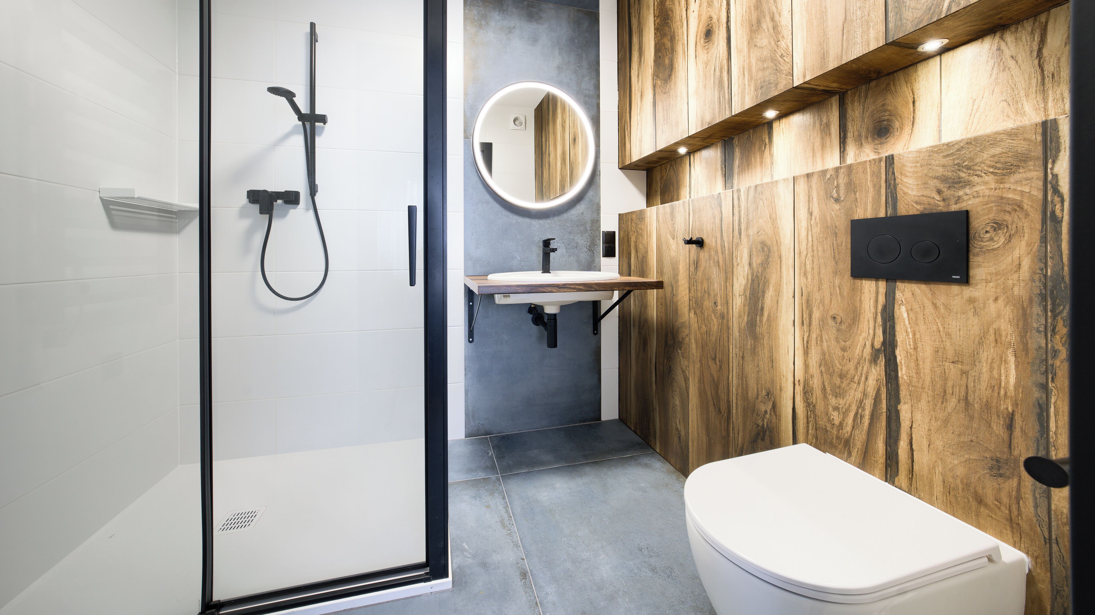 Un baño moderno con cabina de ducha de vidrio, tocador de madera, espejo redondo y inodoro blanco.