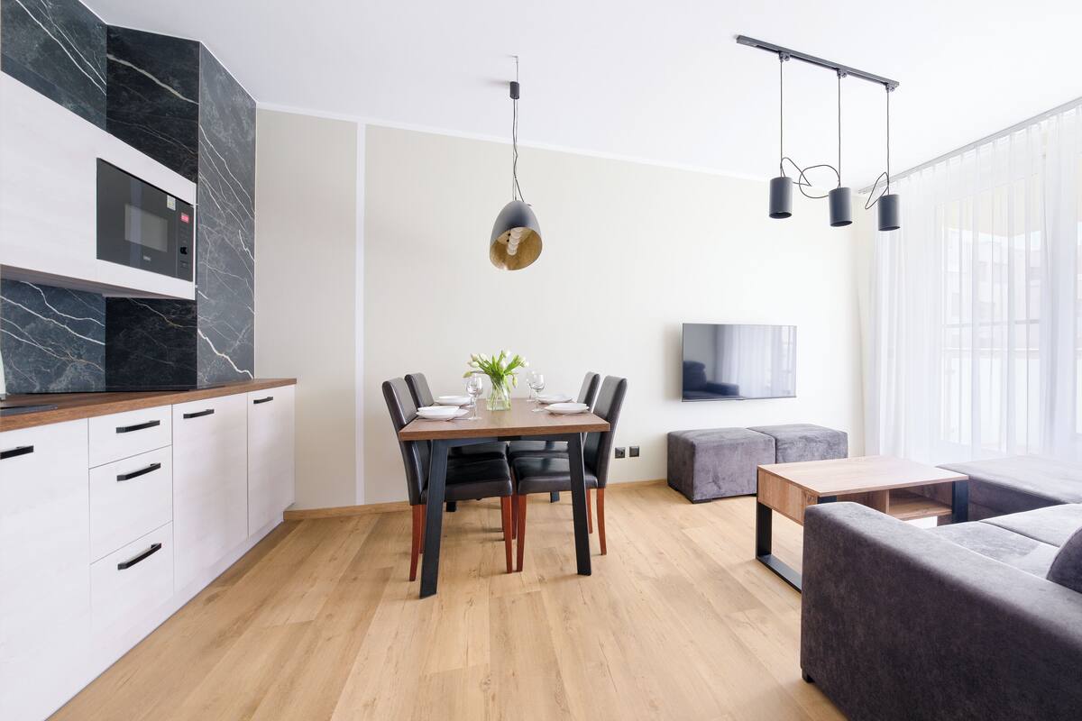 A modern kitchen with a marble backsplash, a dining area with a table and chairs, a grey sofa, and a TV mounted on the wall.