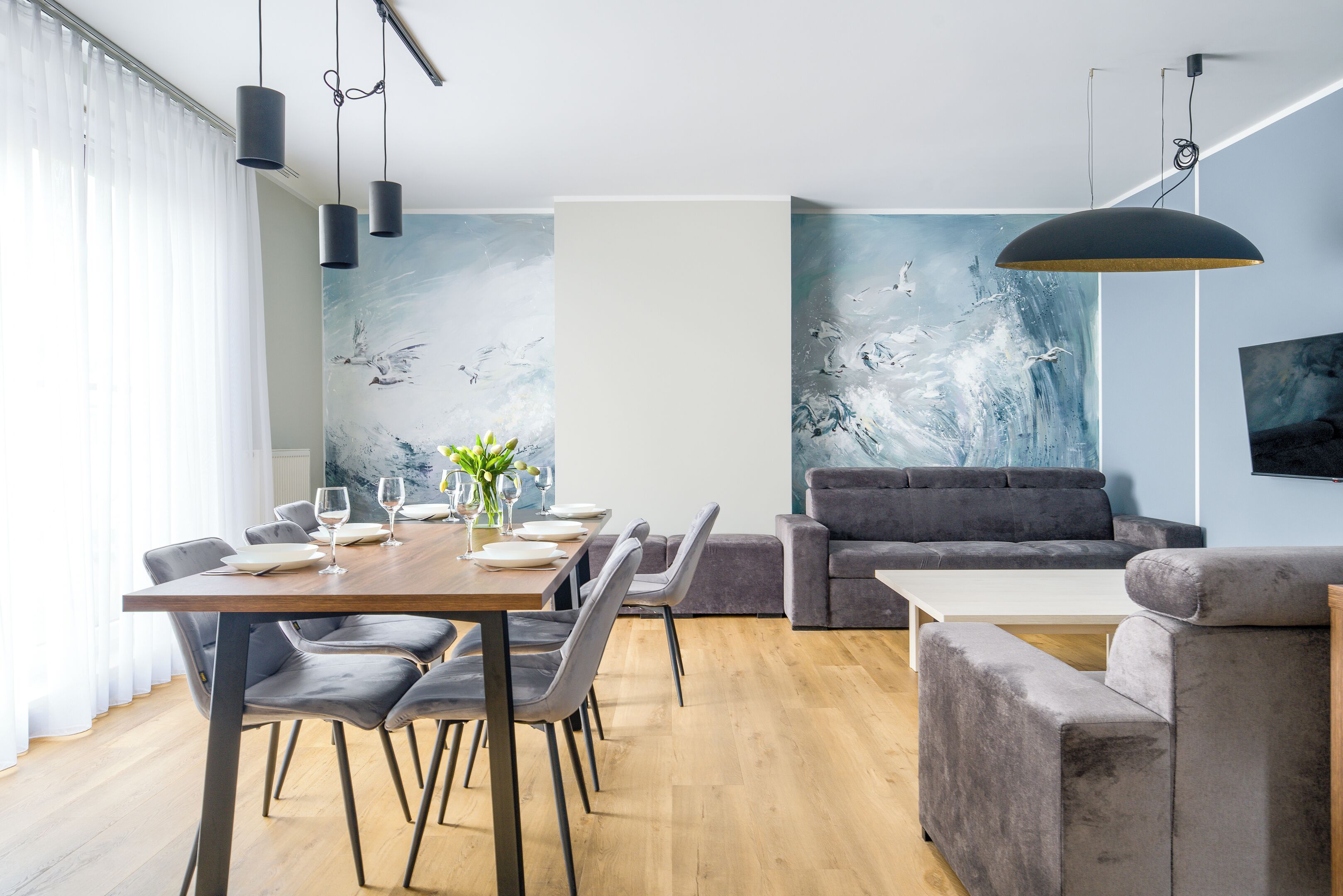 Een moderne woonkamer met een eethoek, met een houten tafel gedekt voor zes personen, grijze banken en abstracte wanddecoratie.