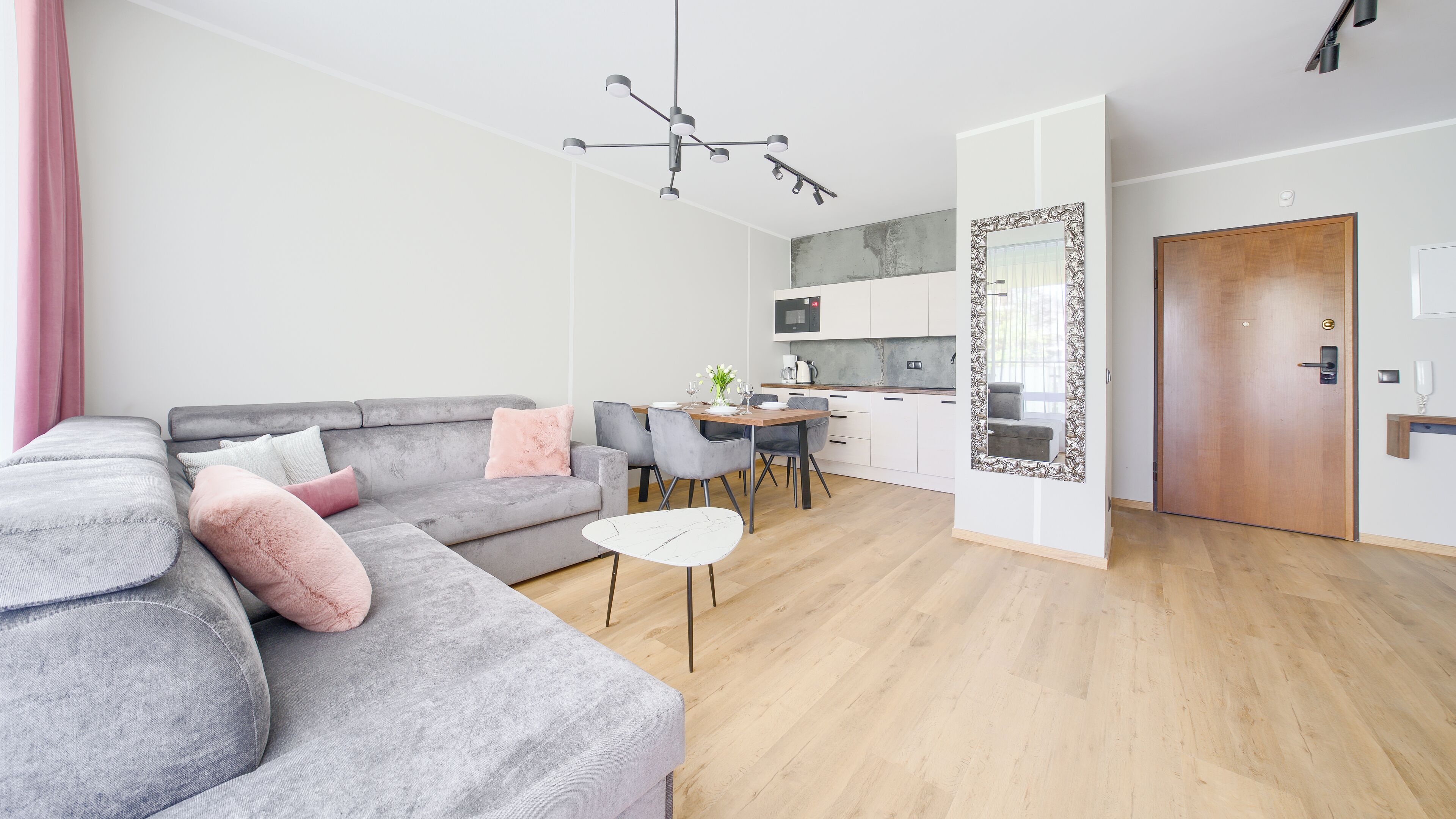 A modern living room with a grey sectional sofa, a small round coffee table, and a dining area with a wooden table and chairs. A kitchen with white cabinets and a large mirror is visible in the background.