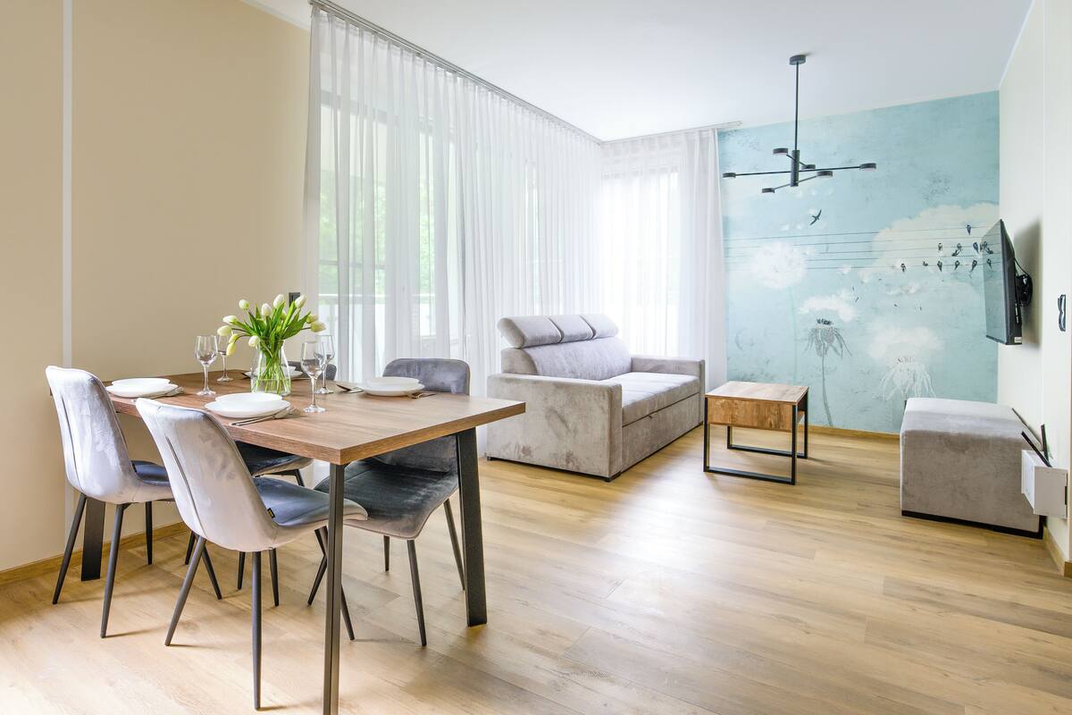 A modern living room with a dining area, featuring a wooden dining table, grey upholstered chairs, a grey sofa, and a small wooden side table.