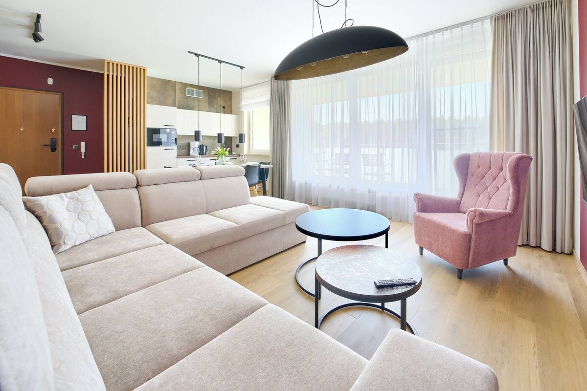A modern living room with a sofa, armchair, and coffee table, a kitchen in the background, and a large window with curtains.