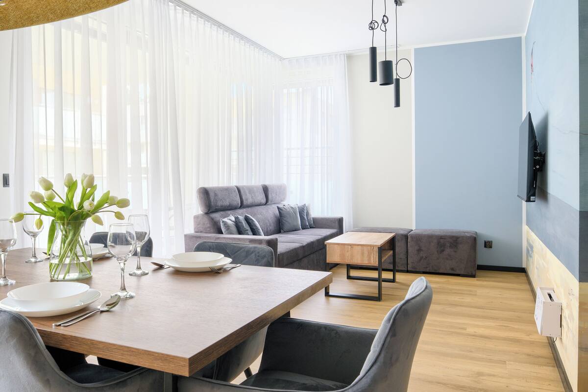 A modern living room with a dining area, featuring a wooden dining table, grey upholstered furniture, and a large window with sheer curtains.