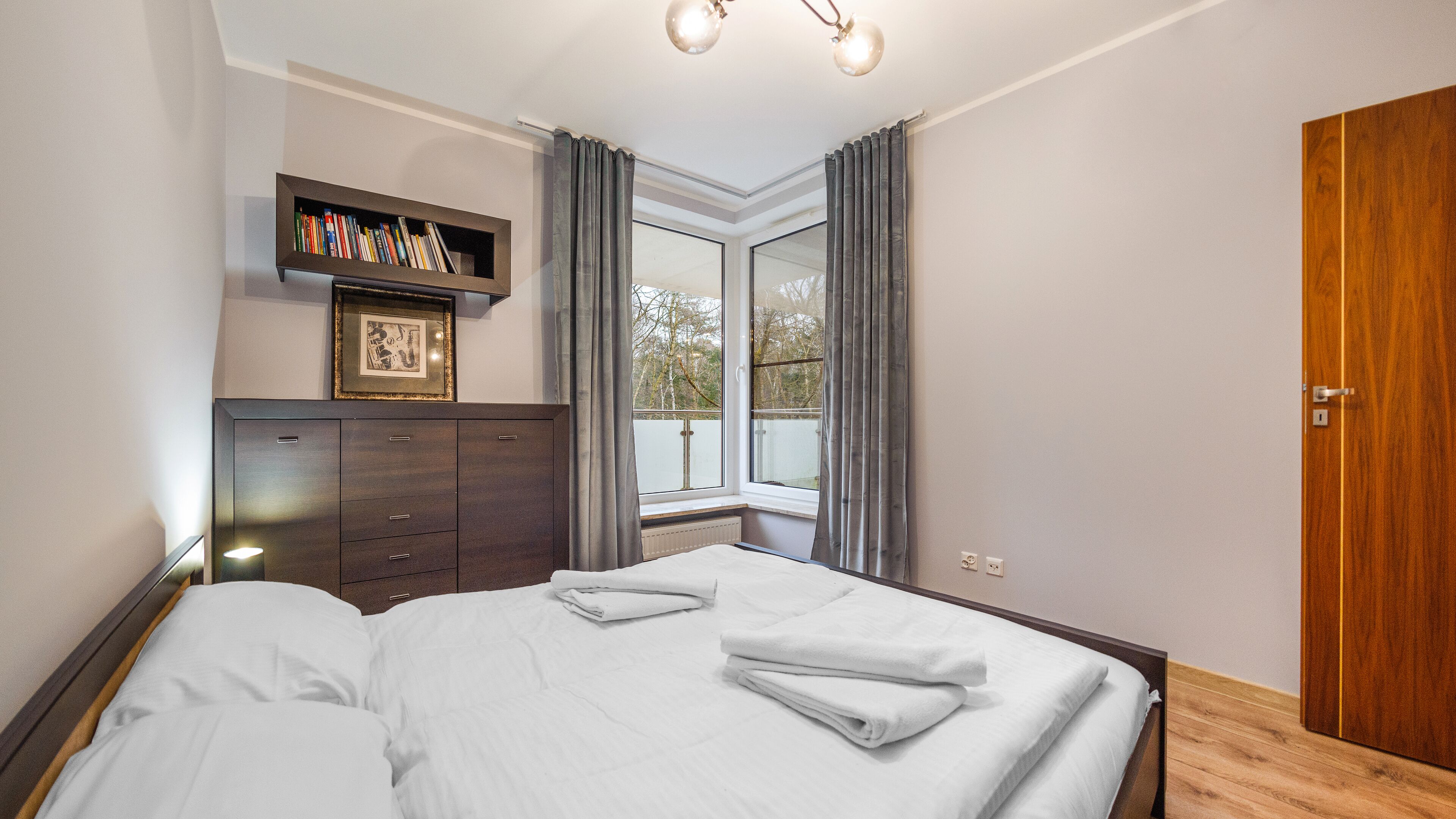 A bedroom with a large bed, a bedside table, a wardrobe, a window with curtains, and a bookshelf with books.