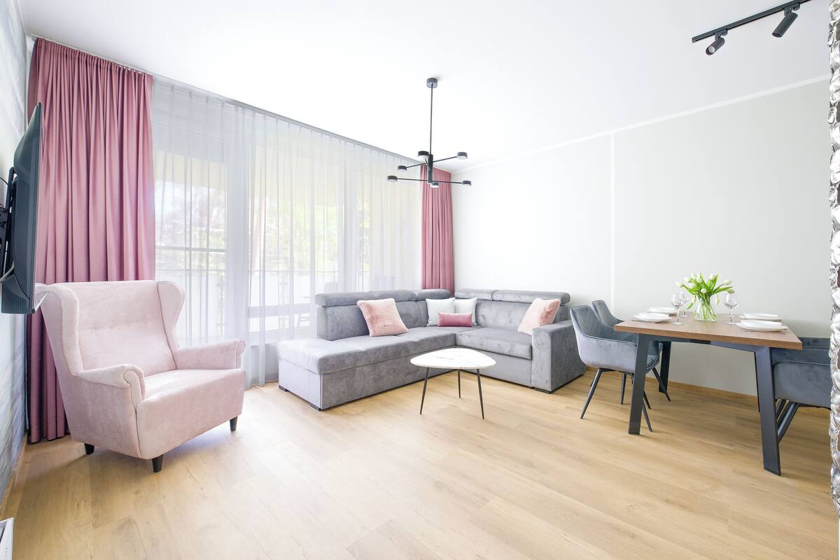 A modern living room with a grey sofa, a pink armchair, a wooden coffee table, and a dining table with a vase of flowers.