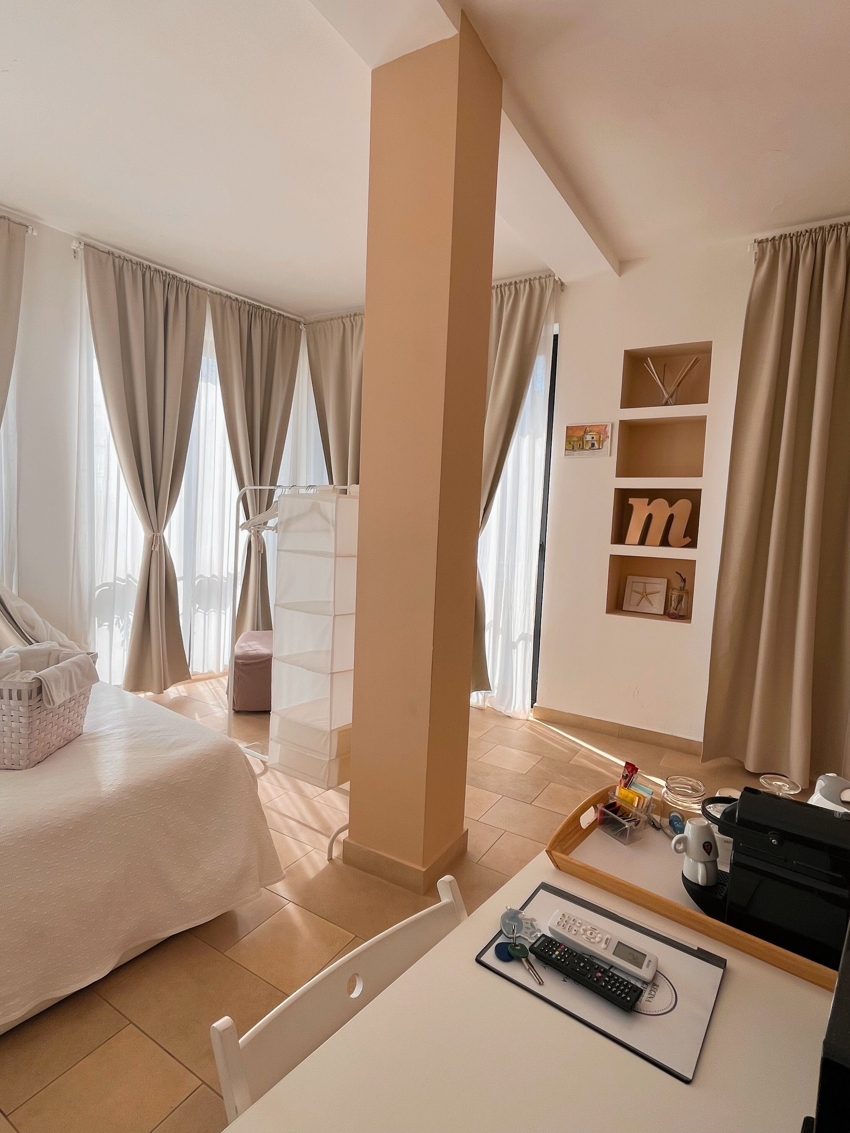 Ein Hotelzimmer mit Bett, Schreibtisch, Rechner, Schlüsseln und einer Tasse. Vorhänge, ein Regal mit Dekoration und ein Sessel sind ebenfalls vorhanden.