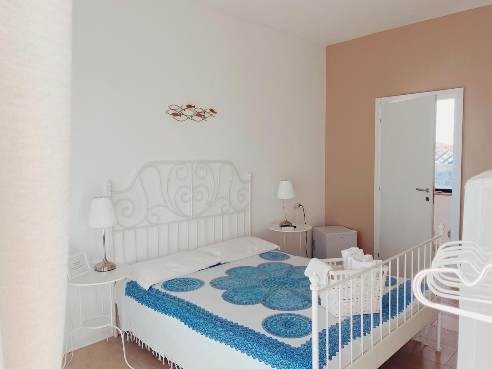 Un dormitorio con una cama blanca, dos mesitas de noche, un tocador, una cuna blanca y una puerta.