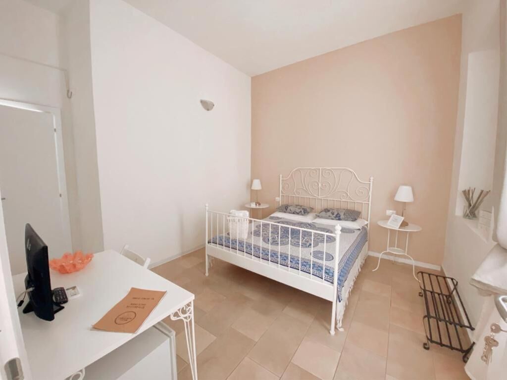 A bedroom with a white metal bed frame, a small bedside table with a lamp, a desk with a computer and a chair, and a small cart.