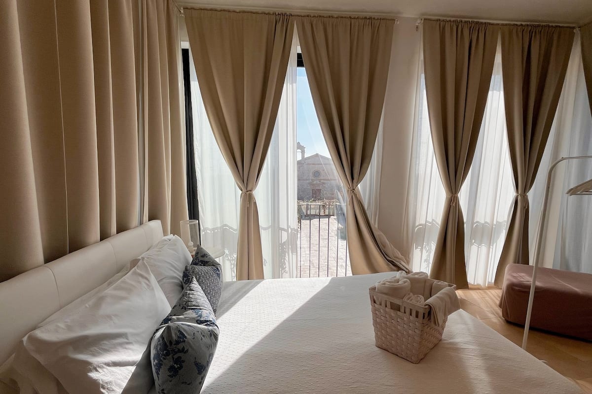 Hotelkamer met een groot bed, beige gordijnen, uitzicht op een gebouw en een klein tafeltje met een mandje.