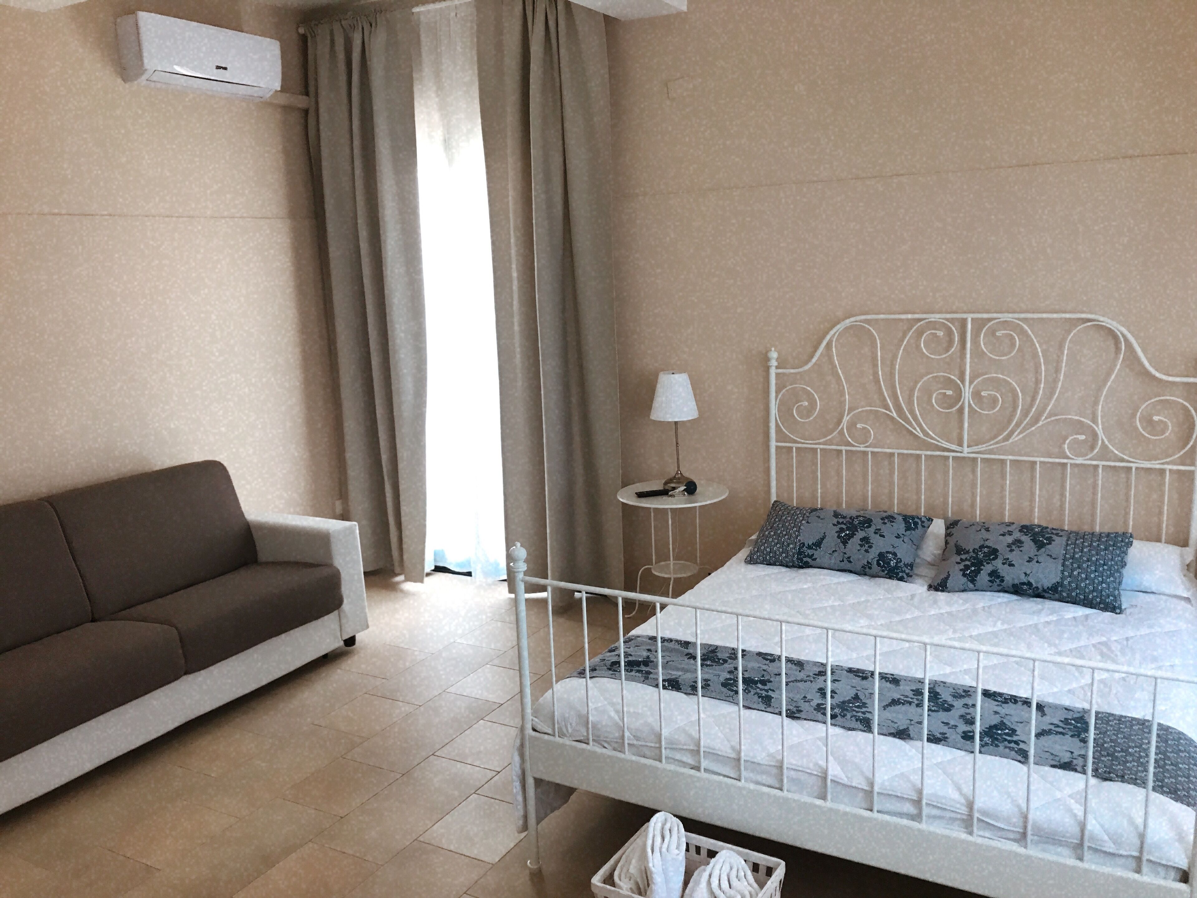 A bedroom with a white metal bed frame, a brown sofa, a small bedside table with a lamp, and a window with curtains.