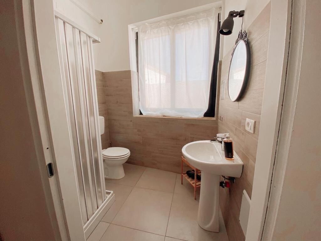 A compact bathroom with a toilet, a pedestal sink, a window with a curtain, and a mirror on the wall.