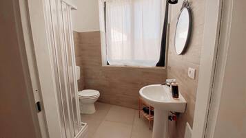 A compact bathroom with a toilet, a pedestal sink, a window with a curtain, and a mirror on the wall.