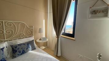 Ein Hotelzimmer mit einem Bett, einem Nachttisch, einer Lampe, einem Fenster mit Vorhängen und einem an der Wand befestigten Regal mit Dekoration.