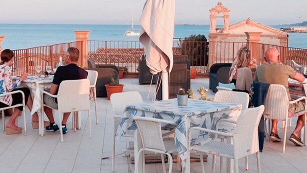 Un espace de restauration en plein air avec des chaises et des tables en osier blanc, un grand parasol, et une vue sur la mer et une arche historique en arrière-plan.