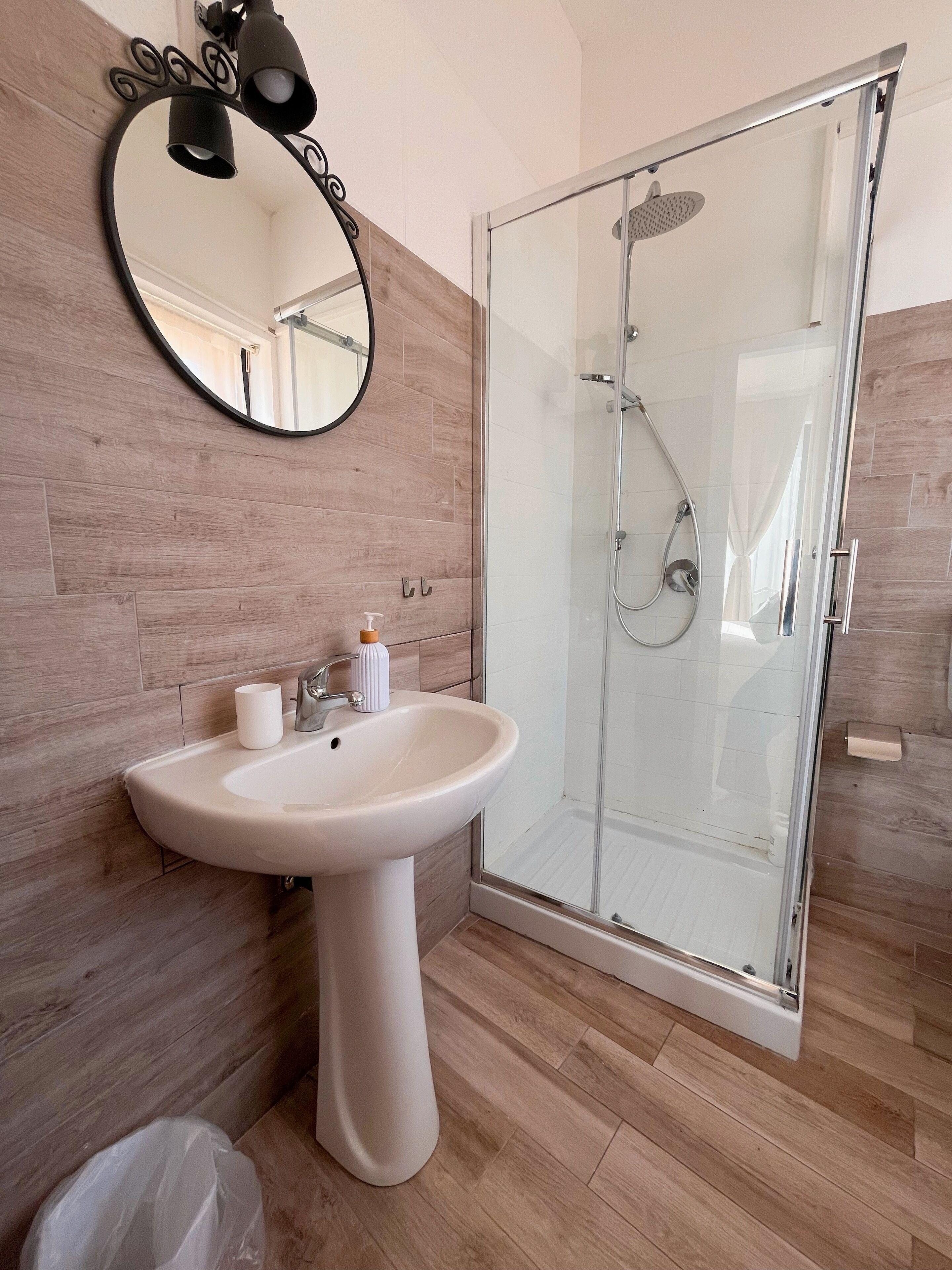 Une salle de bain avec un lavabo sur pied blanc, un miroir rond et une douche à parois de verre.