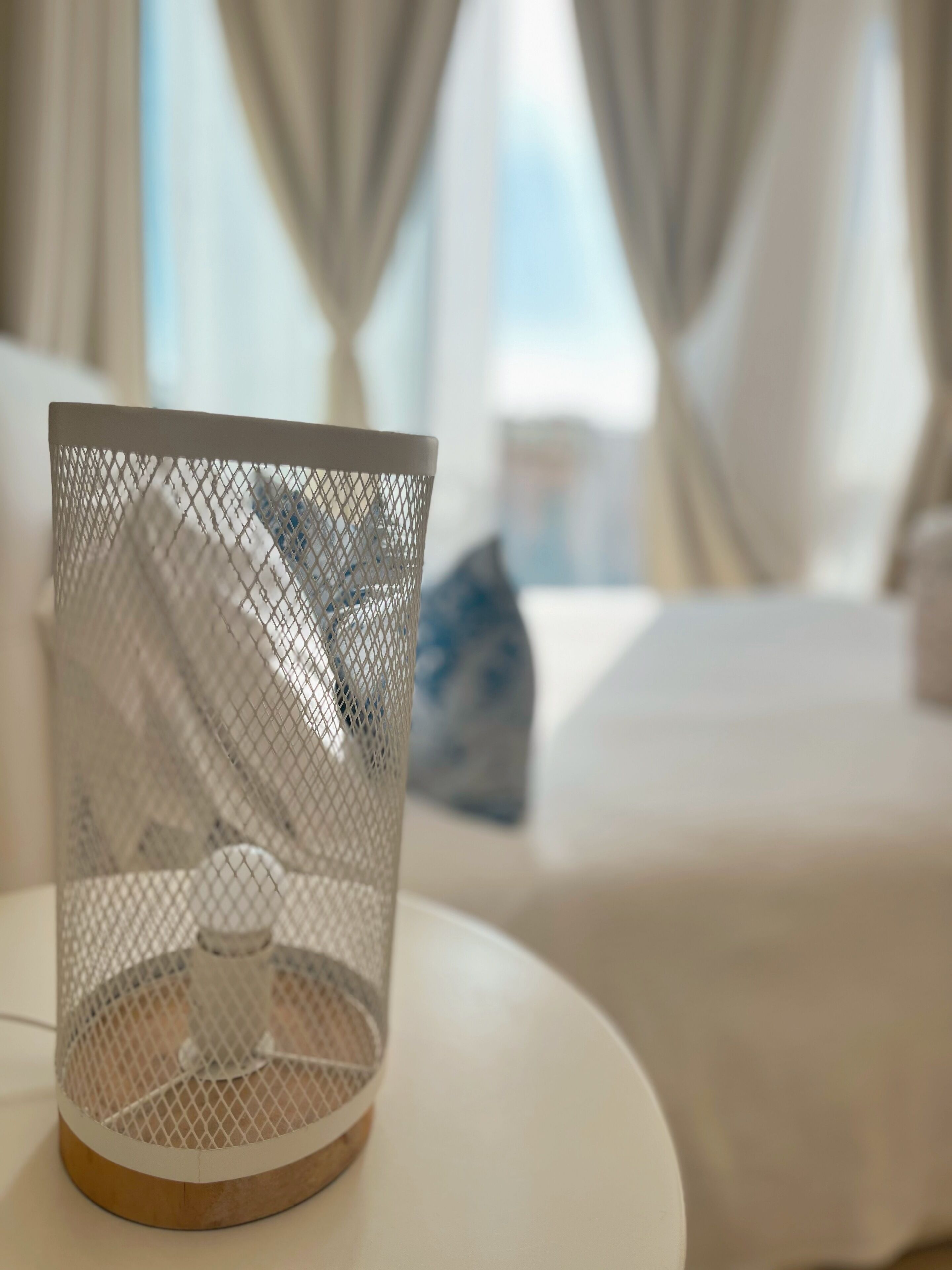 A white mesh candle holder on a round table in a hotel room with a bed and curtains in the background.