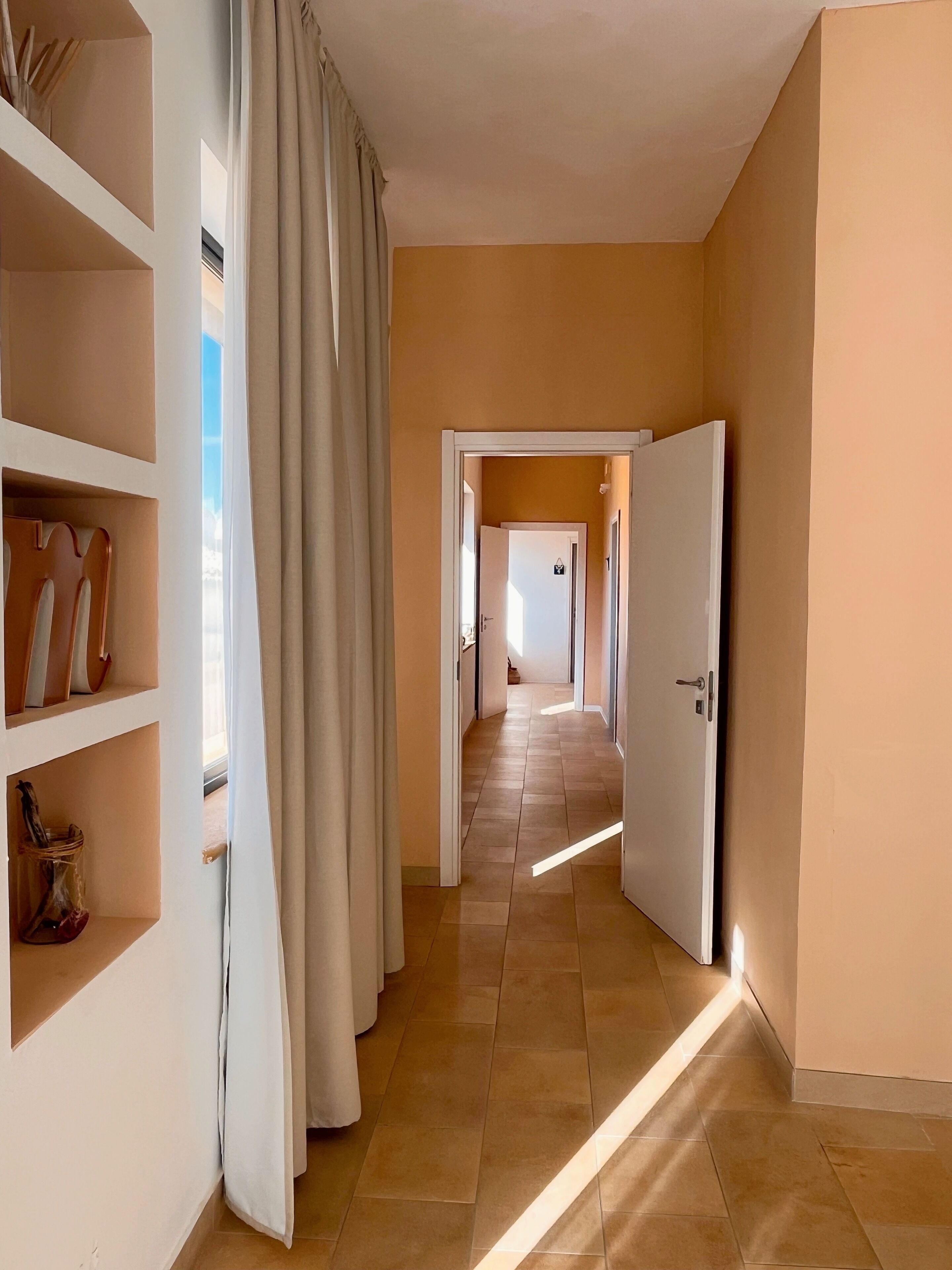 Un pasillo con suelo de baldosas, una puerta blanca a la derecha y una estantería con libros y objetos decorativos a la izquierda.