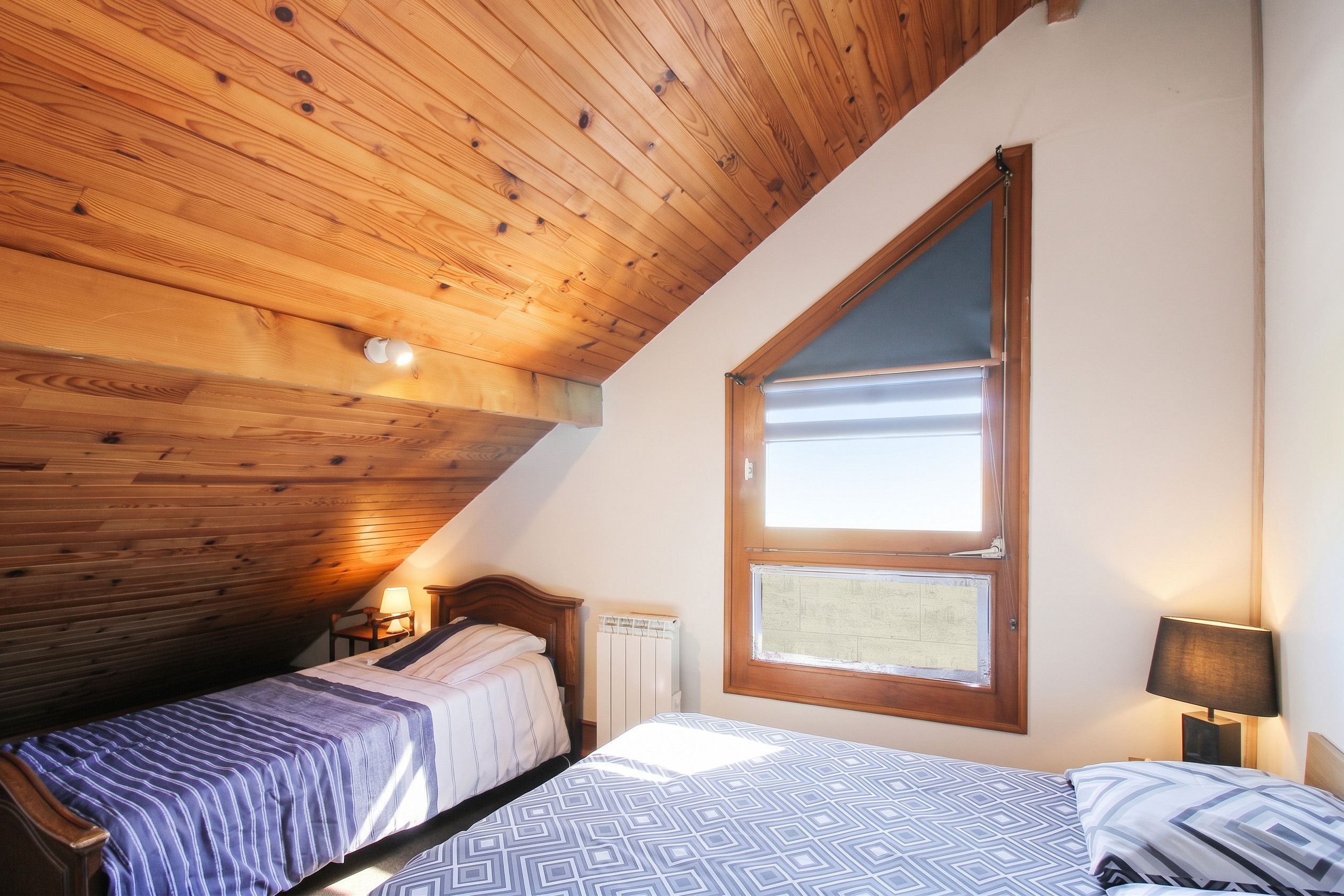 Une chambre avec un plafond en pente, une grande fenêtre, deux lits, une table de chevet et une lampe.