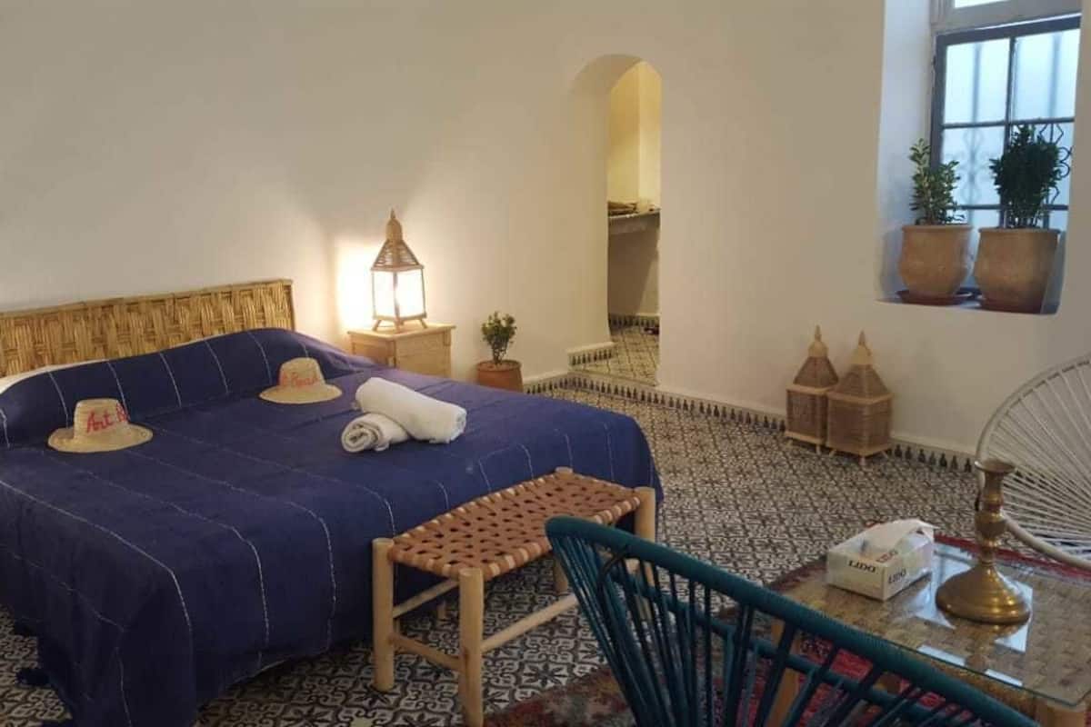 A bedroom with a bed, bedside lamps, a small table, a chair, a mirror, and a window with plants.