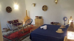 A room with a bed, two chairs, a small table, a lamp, and decorative items on the wall.