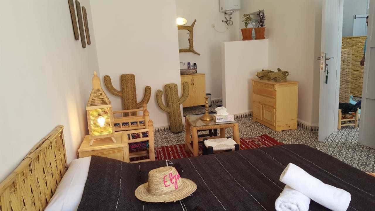 A cozy room with a bed, a small table, a cactus decoration, a wooden cabinet, and a doorway leading to another room.