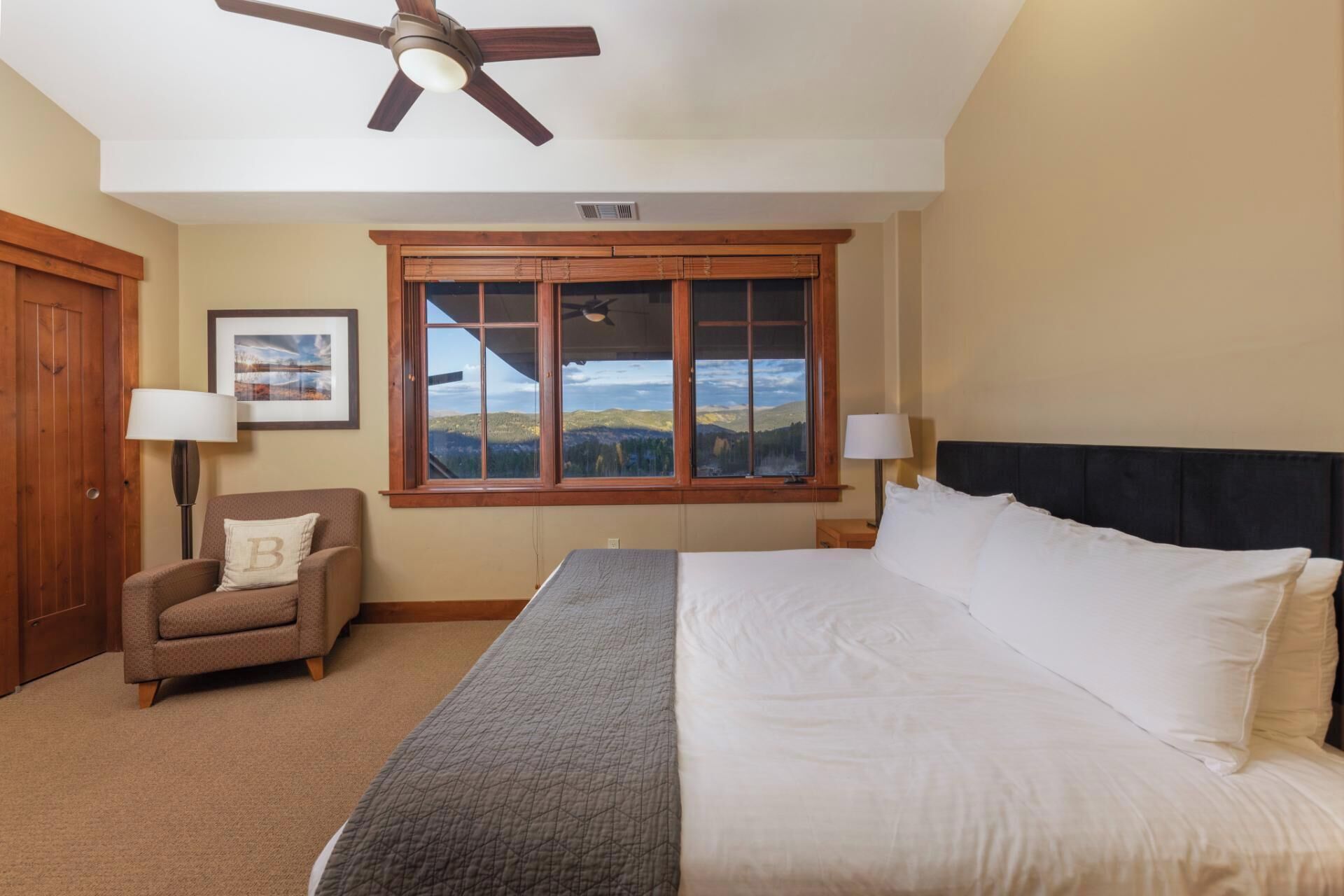A hotel room with a bed, two bedside lamps, a chair, a ceiling fan, a window with a view, and a framed picture on the wall.
